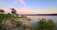 the sun is setting over the water and rocks on the shore with trees in the foreground