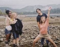 four people standing on rocks in front of water and mountains with their arms up to the sky