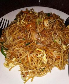 a white plate topped with fried noodles and vegetables next to a fork on top of a table