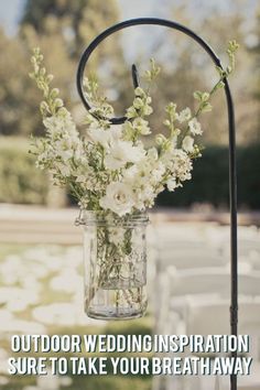 a mason jar with white flowers is hanging from a metal stand at an outdoor ceremony