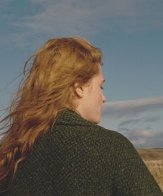 a woman with long red hair standing in a field looking off into the distance while wearing a green sweater