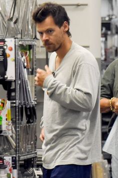 a man in grey shirt standing next to a shelf filled with items and looking at the camera