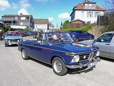 an old blue convertible car driving down a street with other cars parked on the side of the road