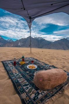 a blanket on the beach with food and drinks in front of it under an awning