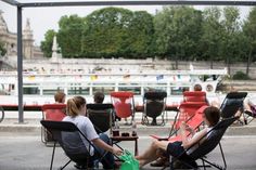 two people sitting on lawn chairs in front of a river with boats and buildings behind them