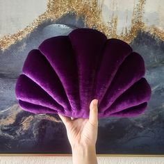 a hand holding up a purple pillow in front of a painting