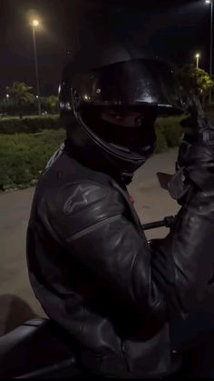 a man wearing a black leather jacket and helmet riding a motorcycle at night with his hand on the handlebars