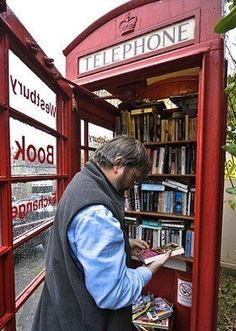 a man standing in a red phone booth with books on the shelves and looking at his cell phone