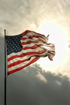 an american flag is flying in the wind on a cloudy day with sun behind it