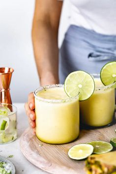 two margaritas with lime wedges on a wooden tray next to copper shakers