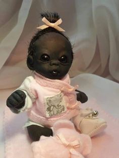 a black baby doll sitting on top of a pink blanket wearing a sweater and booties