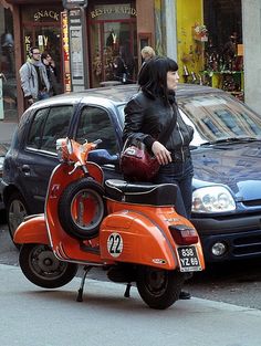 an orange scooter is parked on the side of the road with a woman standing next to it