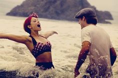 a woman in a bathing suit standing in the water next to a man wearing a hat