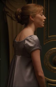 a woman standing in front of a door wearing a gray dress and gold brooch