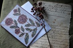 two cards with flowers on them sitting next to an old letter and some berries hanging from it