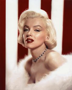 a woman with blonde hair wearing a necklace and white fur stole around her neck, in front of a red striped wall