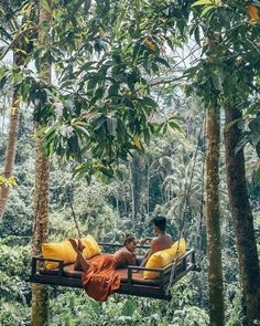 two people sitting on a swing in the middle of some trees with yellow pillows hanging from them