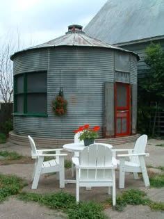 three white chairs around a small table in front of a round building with red doors