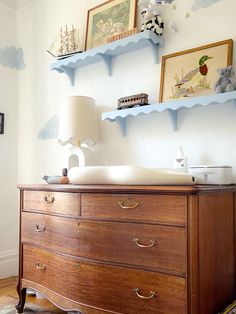 a dresser in a room with shelves and pictures on the wall above it, along with a lamp