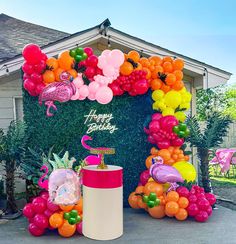 a birthday decoration with balloons and flamingos on the side of a house in front of a pineapple tree