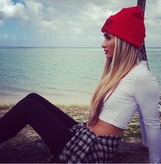 a woman wearing a red beanie sitting on a tree next to the ocean with her legs crossed