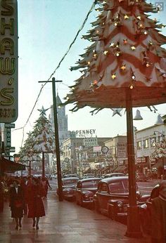 people walking down the sidewalk in front of a christmas tree on a city street with cars parked nearby