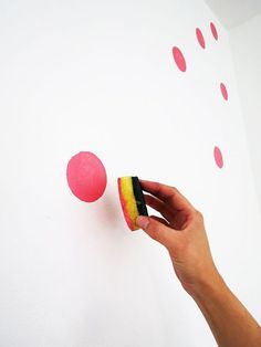 a hand holding a sponge next to a pink and yellow ball on a white wall