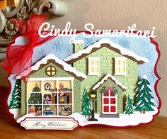 a christmas card with a green house and red bow on it's corner, in front of a holiday tree