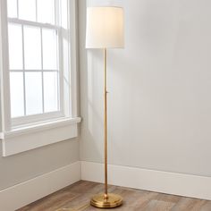 a floor lamp with a white shade on it in front of a window and wooden floor