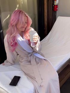 a woman with pink hair is sitting on a bed and holding a cup in her hand