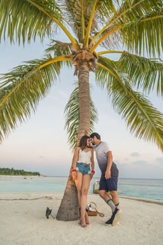 a man and woman standing next to a palm tree on the beach with blue water in the background