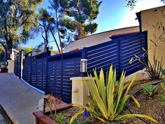 a blue fence next to a sidewalk with plants and trees in front of it on a sunny day