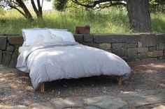 a bed sitting next to a stone wall under a tree with white pillows on it