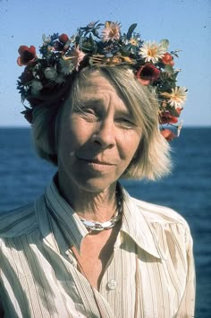 an older woman with a flower crown on her head standing in front of the ocean