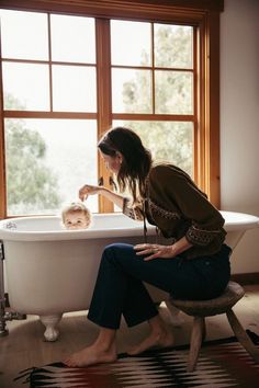 a woman brushing her baby's teeth while sitting in a bathtub next to a window