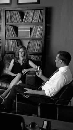 a man and woman are sitting on the couch in front of bookshelves, drinking wine