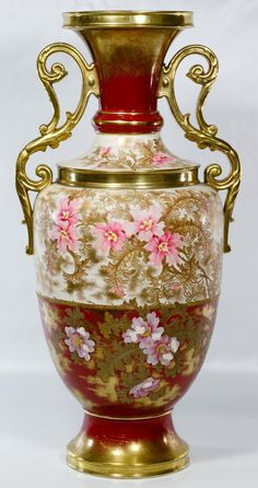 an ornate vase with pink flowers and gold trimmings on the bottom, sitting against a white background