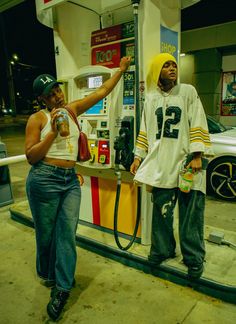 two people standing at a gas pump, one holding a cell phone and the other listening to music