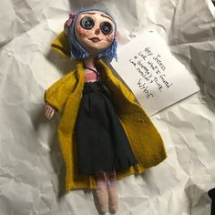 a doll with blue hair wearing a yellow coat and black dress is laying on white paper