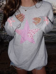 a woman wearing a grey sweatshirt with pink stars on the front and back, standing in front of water