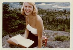 a woman sitting on top of a rock with a book in her hand and trees in the background