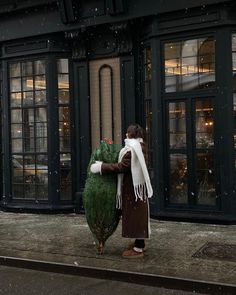 a woman carrying a christmas tree on the sidewalk in front of a building with large windows