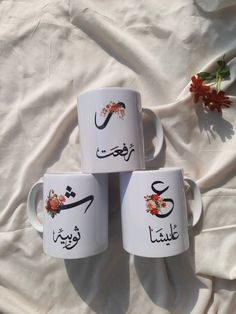 three coffee mugs with arabic writing on them sitting on a white sheet next to flowers