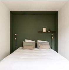 a bed with white sheets and pillows in a green walled room, next to a shelf on the wall