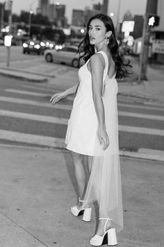 black and white photograph of woman in dress on street corner at night with city lights behind her