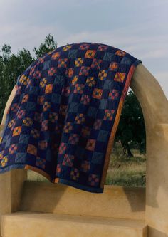 a blue and orange quilt hanging from the side of a stone arch with trees in the background