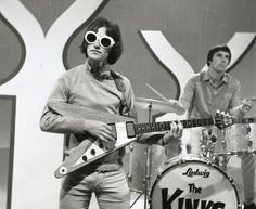 a man with sunglasses on holding a guitar and standing next to another man in front of him