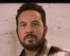 a man with a beard and plaid shirt looking at the camera while standing in front of a white wall