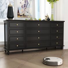 a roomba sits on the floor in front of a black dresser with gold knobs