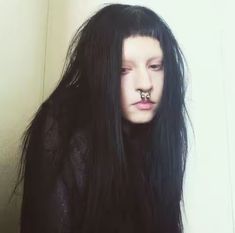 a woman with long black hair and piercings on her nose is standing in front of a white wall